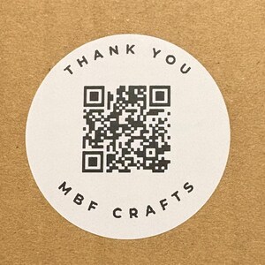 May include: A brown cardboard box with a white circular sticker. The sticker features a QR code, surrounded by the words "THANK YOU" and "MBF CRAFTS". The box is square and has a simple, clean design.