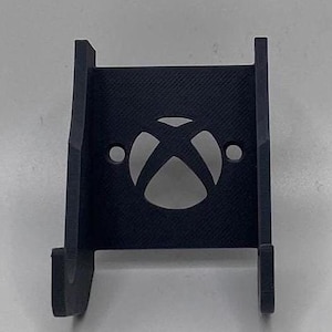 May include: Black plastic wall mount for a video game console. The mount has a cut-out in the shape of the Xbox logo.