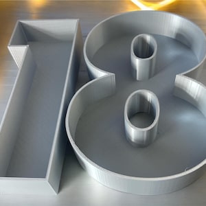 May include: Two grey, 3D printed numbers, 18, are displayed on a silver surface. The numbers are hollow and have a smooth, slightly textured finish.