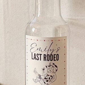 May include: Clear glass bottle with a gold cap and a label that reads "Emily's Last Rodeo" with a cartoon illustration of a woman in a cowboy hat. The date "MAY 20, 2026" is printed below.