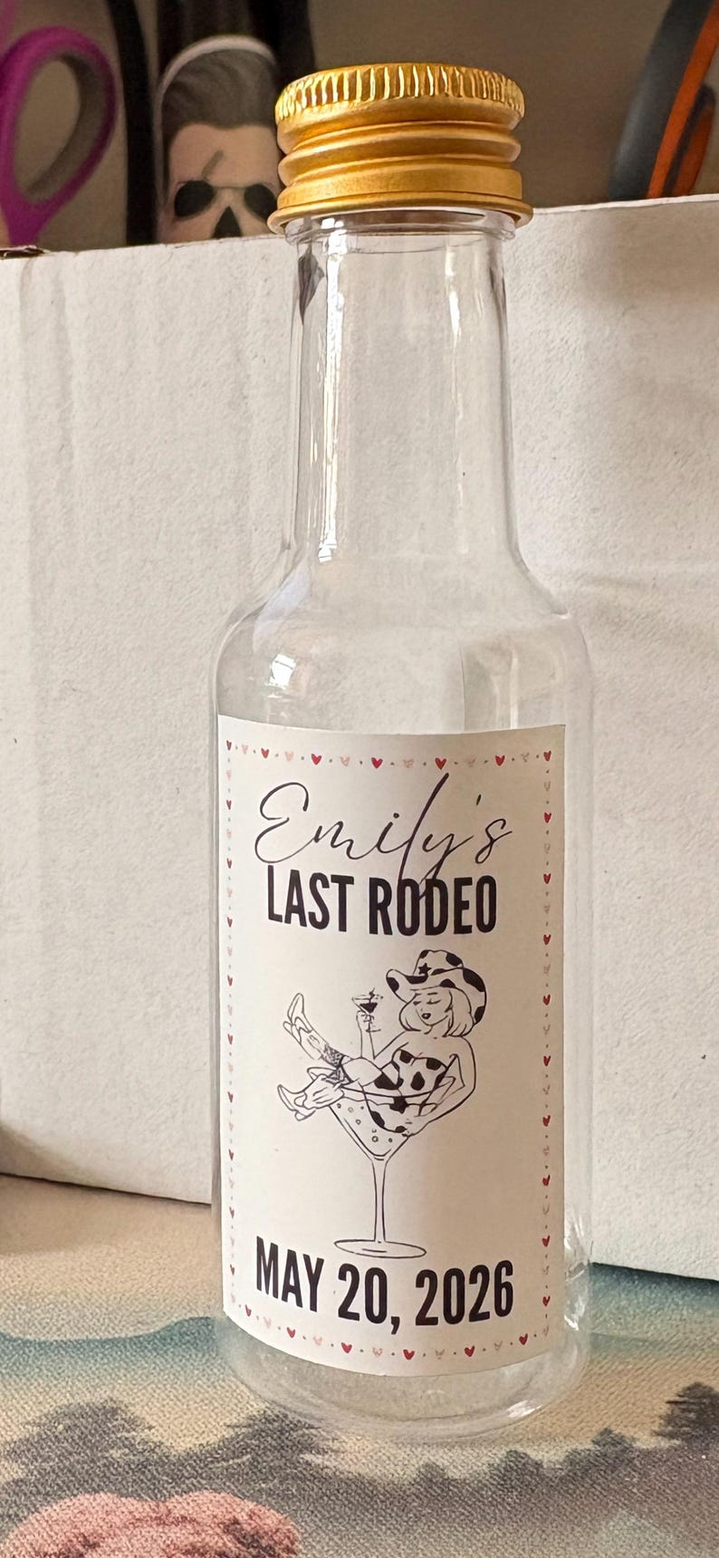 May include: Clear glass bottle with a gold cap and a label that reads "Emily's Last Rodeo" with a cartoon illustration of a woman in a cowboy hat. The date "MAY 20, 2026" is printed below.