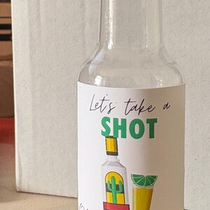 May include: Clear glass bottle with a gold cap. The label reads "Let's take a SHOT" with illustrations of a tequila bottle, a shot glass, and a lime wedge. The text "Olivia's tying the KNOT" is also on the label.