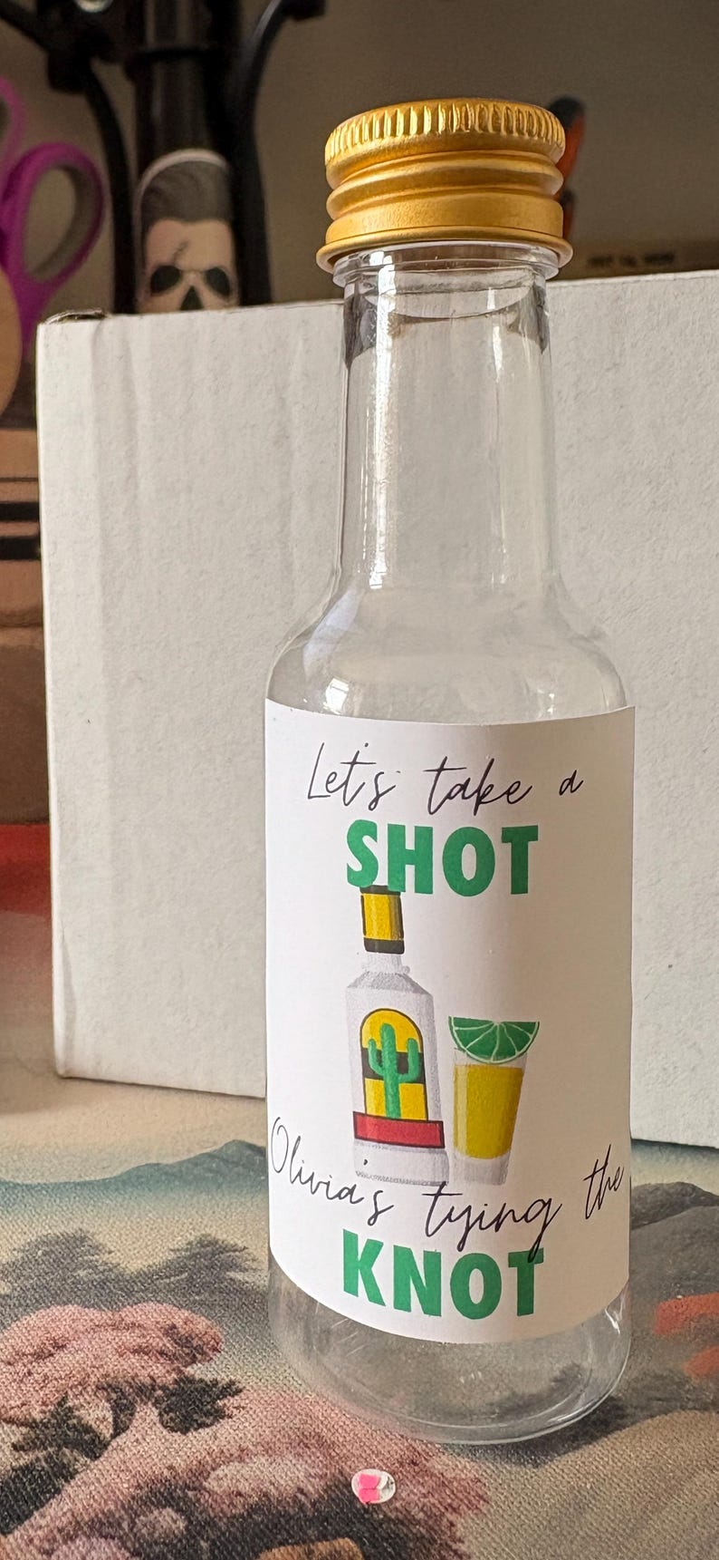 May include: Clear glass bottle with a gold cap. The label reads "Let's take a SHOT" with illustrations of a tequila bottle, a shot glass, and a lime wedge. The text "Olivia's tying the KNOT" is also on the label.