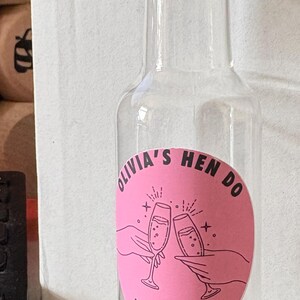 May include: A clear glass bottle with a gold-coloured cap, displaying a pink label. The label reads "Olivia's Hen Do June 2026" and features two champagne glasses. The bottle is designed for a celebration.
