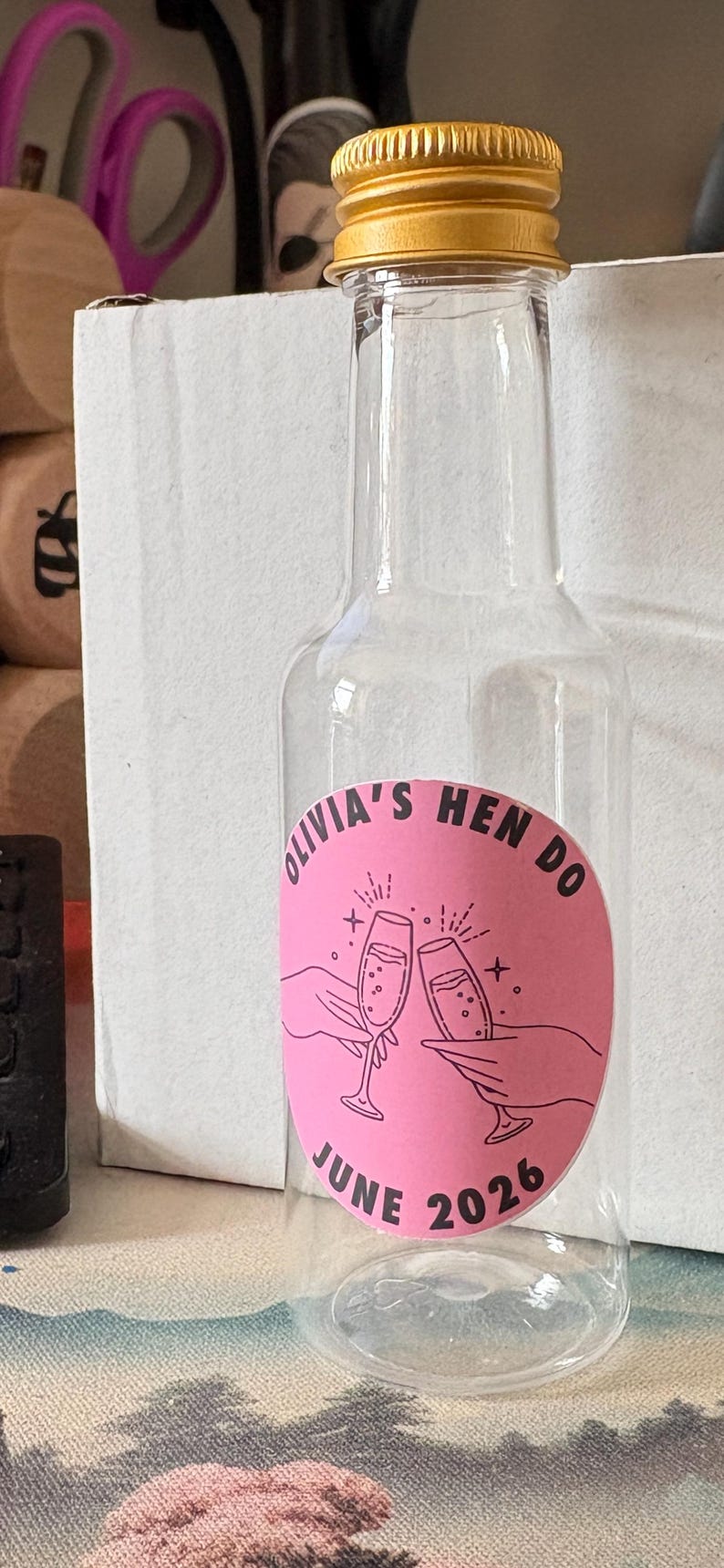 May include: A clear glass bottle with a gold-coloured cap, displaying a pink label. The label reads "Olivia's Hen Do June 2026" and features two champagne glasses. The bottle is designed for a celebration.