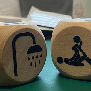 May include: Two wooden dice with black illustrations. One die shows a couch and a shower, the other depicts sexual positions. The dice are light brown with a smooth finish. The dice are on a green surface.