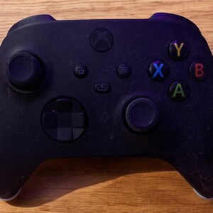 May include: A black Xbox controller with a textured surface. The controller has a directional pad, four face buttons labelled A, B, X, and Y, and two analogue sticks.
