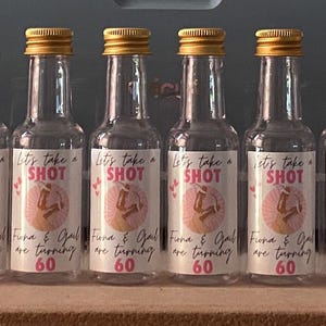 May include: A collection of clear glass shot bottles with gold-coloured caps. Each bottle features a white label with the word "SHOT" in pink, alongside other text and a pink graphic. The bottles are arranged on a wooden surface.
