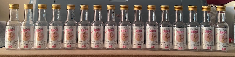 May include: A collection of clear glass shot bottles with gold-coloured caps. Each bottle features a white label with the word "SHOT" in pink, alongside other text and a pink graphic. The bottles are arranged on a wooden surface.