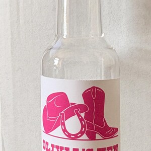 May include: A clear glass bottle with a gold-coloured cap. The bottle features a label with a pink cowboy hat, boot, and horseshoe graphic, and the text "OLIVIA'S HEN".