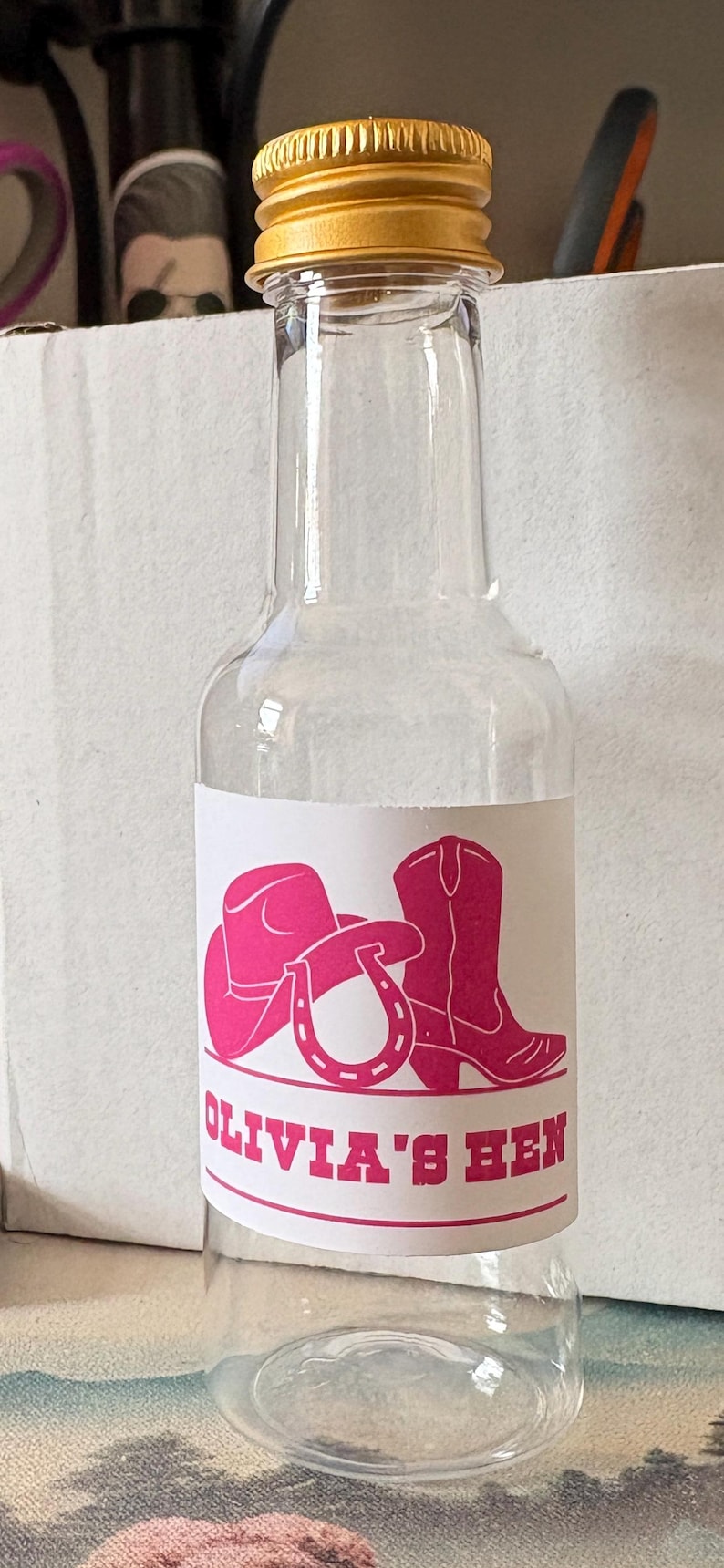May include: A clear glass bottle with a gold-coloured cap. The bottle features a label with a pink cowboy hat, boot, and horseshoe graphic, and the text "OLIVIA'S HEN".