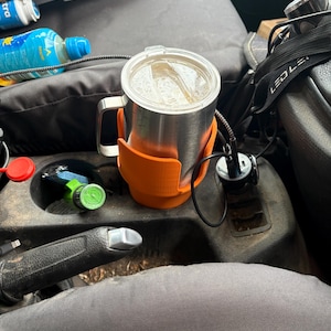 May include: A stainless steel travel mug with a clear plastic lid and a handle, held securely within a bright orange, custom-fitted 3D-printed cup holder adapter, positioned inside a vehicle centre console cup holder area.