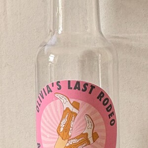 May include: A clear plastic bottle with a gold-coloured cap. The pink label reads "Olivia's Last Rodeo" with a cowboy boot graphic and the date 27 August 2026. Likely a party favour bottle.