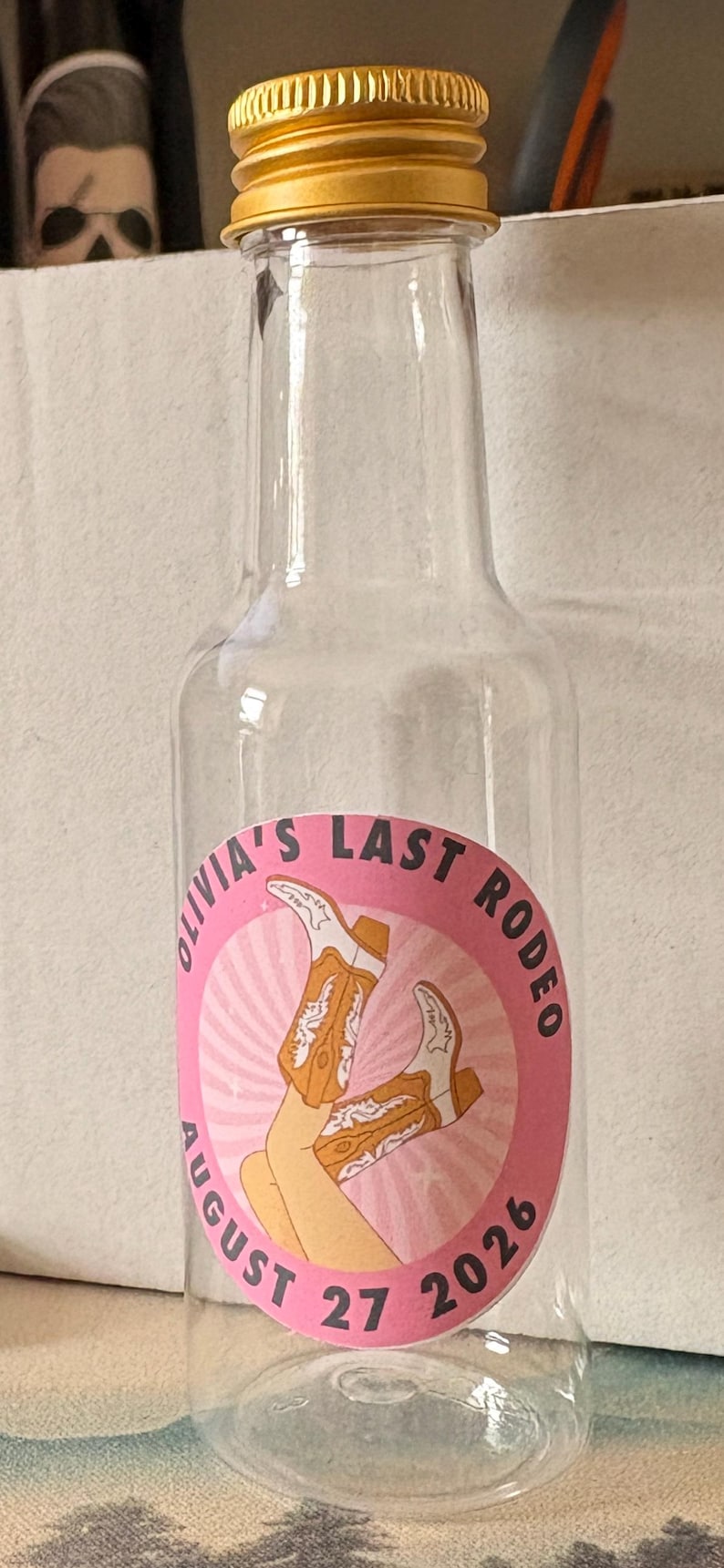 May include: A clear plastic bottle with a gold-coloured cap. The pink label reads "Olivia's Last Rodeo" with a cowboy boot graphic and the date 27 August 2026. Likely a party favour bottle.