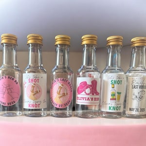 May include: Six small glass bottles with gold caps, each with a label. The labels feature text and graphics, including the phrases "Let's take a SHOT" and "Olivia's Hen Do." The bottles are likely for a party or celebration.