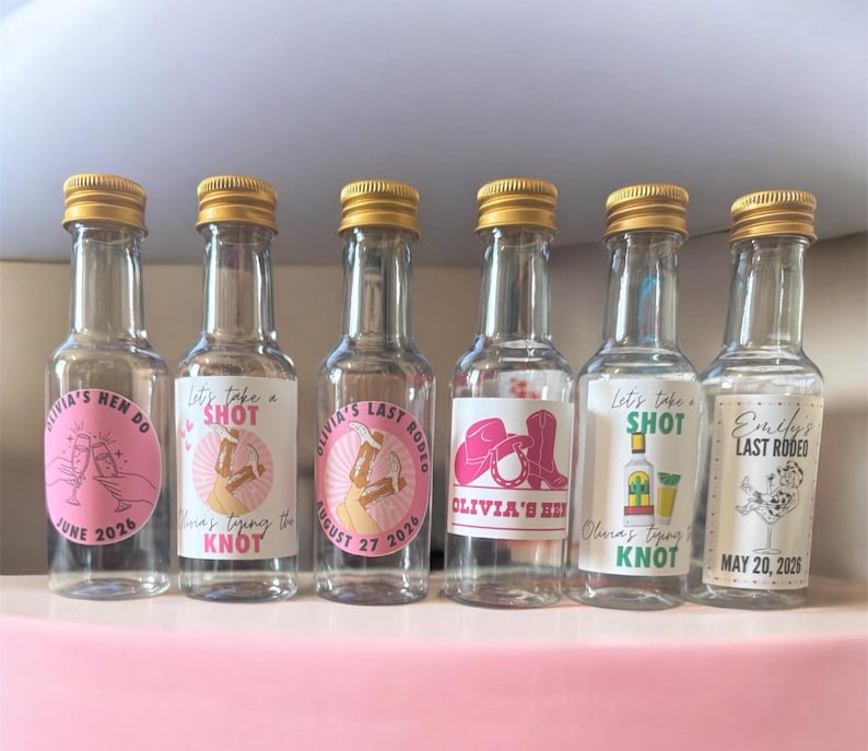 May include: Six small glass bottles with gold caps, each with a label. The labels feature text and graphics, including the phrases "Let's take a SHOT" and "Olivia's Hen Do." The bottles are likely for a party or celebration.
