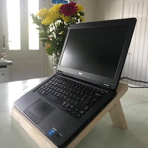 May include: A black Dell laptop computer is placed on a wooden laptop stand. The stand is made of light wood and has a simple design. The laptop is open and the screen is black.