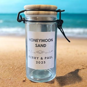 May include: A glass jar with a wooden lid and metal clasp, labelled "Honeymoon Sand", with the names Kerry & Paul and the year 2025. The jar is on a sandy beach with the sea in the background.