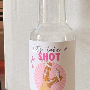 May include: Clear glass bottle with a gold cap, featuring a label with the text "Let's take a SHOT" and "Olivia's tying the KNOT" in pink. The label includes a graphic of cowboy boots.