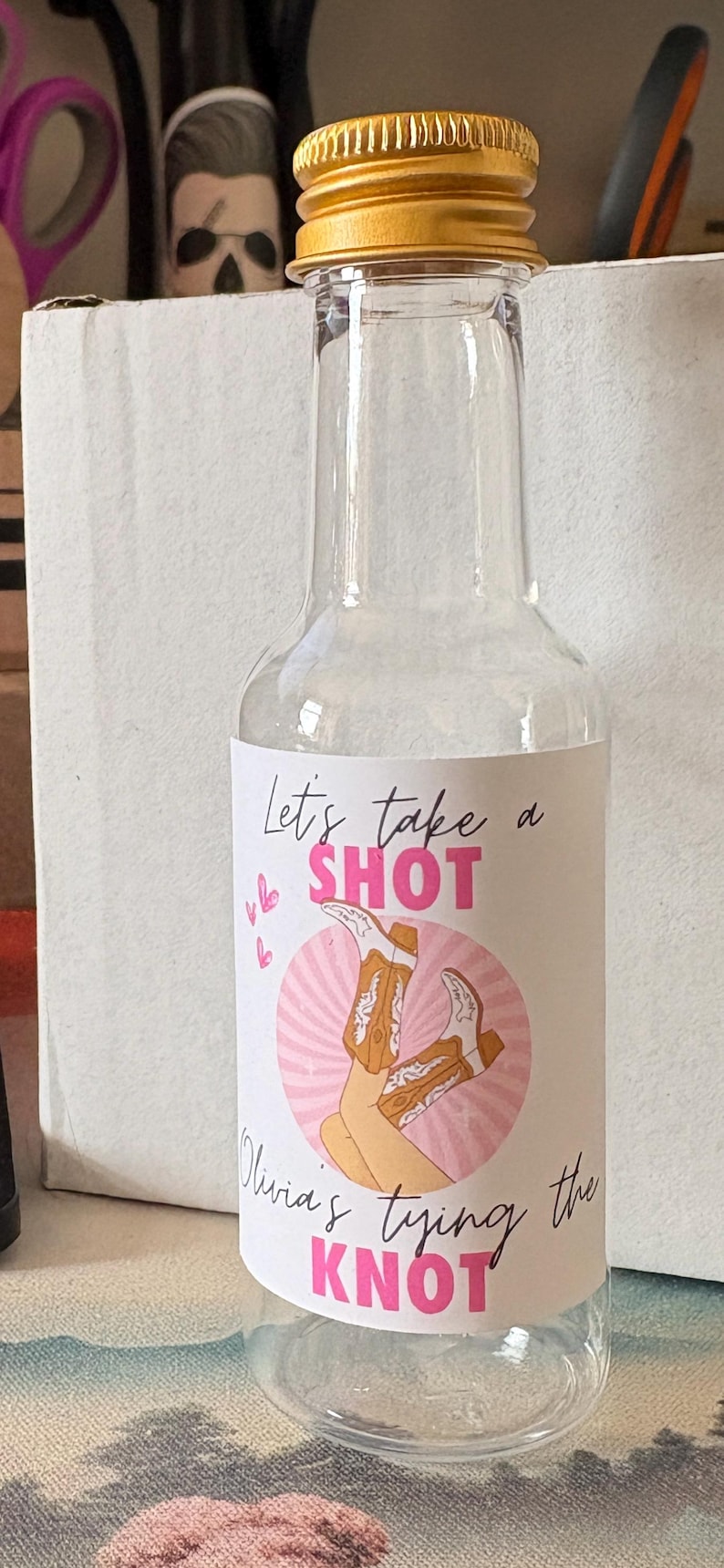 May include: Clear glass bottle with a gold cap, featuring a label with the text "Let's take a SHOT" and "Olivia's tying the KNOT" in pink. The label includes a graphic of cowboy boots.