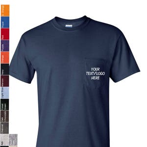 May include: A navy blue t-shirt with a pocket on the left side. The pocket has white text that says "YOUR TEXT/LOGO HERE".