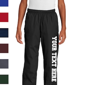 May include: Black athletic pants with a white 'YOUR TEXT HERE' printed down the right leg. The pants have an elastic waistband and a drawstring.