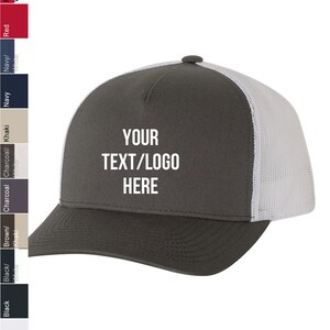 May include: A gray and white trucker hat with a mesh back. The front of the hat has the text "YOUR TEXT/LOGO HERE" embroidered in white.