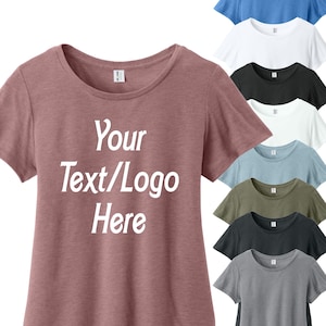 May include: A collection of short-sleeved t-shirts in various colors, including dusty rose, white, black, blue, and gray. The front of the dusty rose shirt displays the text "Your Text/Logo Here" in white, indicating customization options.