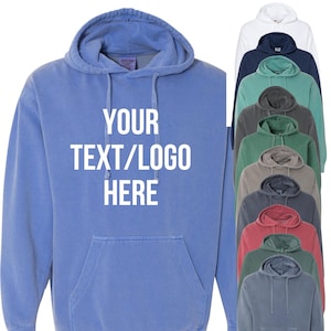 May include: A blue hooded sweatshirt with the text "YOUR TEXT/LOGO HERE" in white. The image also shows a variety of other colored hoodies, including white, navy, green, gray, red, and teal. The sweatshirts have a front pocket and a drawstring hood.