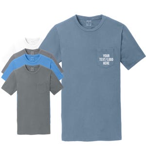 May include: A collection of short-sleeve crew neck t-shirts in various solid colors including blue, gray, and white. Each shirt features a single chest pocket. The blue shirt in the foreground displays the white text YOUR TEXT/LOGO HERE.