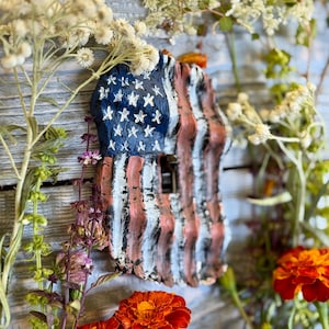 May include: A rustic, hand-painted wooden decoration in the shape of the American flag. The flag features blue, white, and red stripes and stars. The decoration is surrounded by various flowers and greenery, including marigolds.