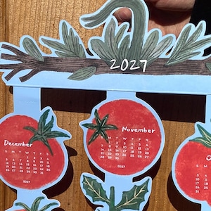 May include: A 2027 calendar with tomato illustrations. The calendar has a light blue background with red tomato-shaped monthly calendars. The months of October, November, and December are visible. The top of the calendar has a branch design with the year 2027.