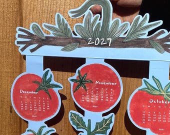 Puede incluir: Un calendario colorido de 2027 con ilustraciones de tomates. El calendario tiene un fondo azul claro con calendarios mensuales rojos en forma de tomate. Se ven los meses de octubre, noviembre y diciembre. La parte superior del calendario muestra el año 2027.