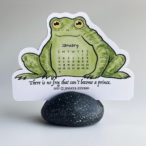 Könnte beinhalten: Eine gestanzte Karte mit einer grünen Froschillustration und einem Kalender für Januar 2027 auf dem Bauch. Der Text lautet "There is no frog that can't become a prince." und "2027 © JENATA STUDIO." Die Karte wird von einem dunkelgrauen, gesprenkelten Stein aufrecht gehalten.