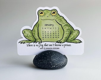 May include: A die-cut card featuring a green frog illustration with a January 2027 calendar on its belly. The text reads, "There is no frog that can't become a prince." and "2027 © JENATA STUDIO." The card is held upright by a dark grey speckled stone.