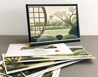 May include: A 2027 desk calendar titled Garden of Quiet Seasons, displayed in a black frame. The calendar features illustrations of a Japanese-style garden with trees, shrubs, and a circular window. Loose calendar pages are stacked below the frame.