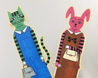 May include: Two die-cut paper bookmarks featuring anthropomorphic animal illustrations. On the left, a green cat with black stripes wears a blue dress and carries a grey handbag. On the right, a pink rabbit wears a brown dress with a black bow and carries a cream handbag.
