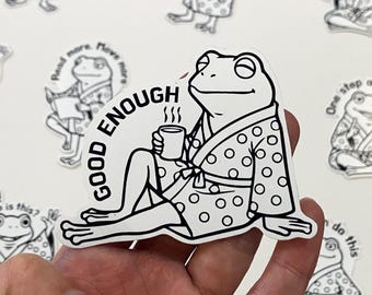 May include: A black and white sticker of a frog in a robe, holding a mug. The text "GOOD ENOUGH" is in a circle around the frog. Other stickers with frogs are visible in the background.