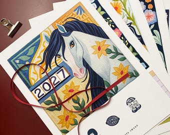 May include: A 2027 calendar page featuring a white horse with a dark mane, surrounded by yellow flowers and green leaves on a textured background. A red ribbon lies across the print, which is stacked with other illustrated pages and a metal clip.