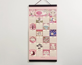 May include: A 2027 wall calendar featuring a pink background and twelve monthly grids. Each month is illustrated with distinct Japanese-inspired motifs including a crane, pagoda, cherry blossoms, Mount Fuji, a frog, koi fish, and rabbits, held by black wooden rails.