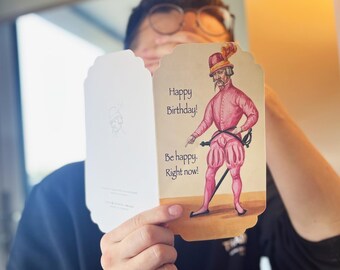 May include: A birthday card featuring a cartoon illustration of a man in pink attire and a feathered hat. The card displays the text "Happy Birthday! Be happy. Right now!" The card is held open, showing the inside.