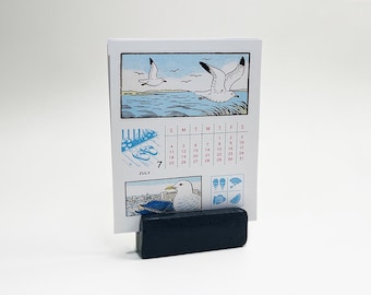May include: A white paper calendar for July featuring illustrations of seagulls, a coastal scene, and summer icons, held upright by a rectangular black textured base. The calendar displays the month name, dates, and small blue graphic prints.