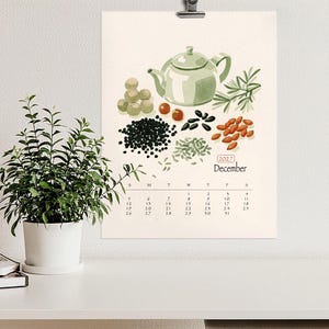 Peut inclure: Une page de calendrier mural pour décembre 2027 illustrée d'une théière vert clair, de brins d'herbes et de diverses graines et baies. La grille du calendrier affiche les jours du mois sous l'illustration sur un fond couleur crème.