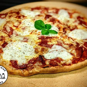 May include: A round pizza with a golden-brown crust, topped with red tomato sauce, melted white mozzarella cheese, slices of pepperoni, and a garnish of fresh green basil leaves. The pizza is placed on a round stone surface with a logo in the corner.