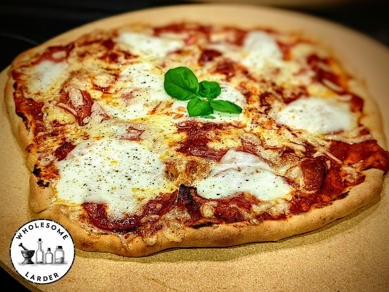 May include: A round pizza with a golden-brown crust, topped with red tomato sauce, melted white mozzarella cheese, slices of pepperoni, and a garnish of fresh green basil leaves. The pizza is placed on a round stone surface with a logo in the corner.