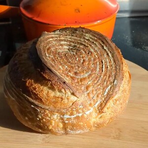 May include: A round loaf of crusty sourdough bread with a spiral flour pattern on top, positioned in front of a bright orange enameled cast iron Dutch oven with a lid. A circular logo in the top left corner reads WHOLESOME LARDER.