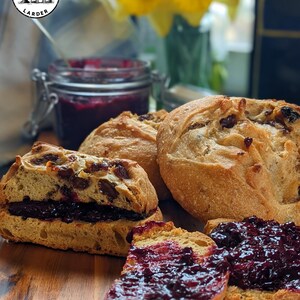 May include: Several round, golden-brown artisan bread rolls containing dark dried fruit, with one roll sliced in half and spread with thick, dark purple berry jam. A glass jar of the same jam sits in the background with a logo reading WHOLESOME LARDER.