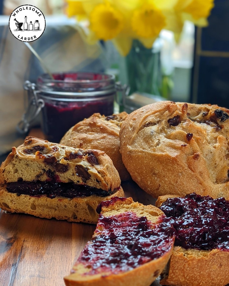 May include: Several round, golden-brown artisan bread rolls containing dark dried fruit, with one roll sliced in half and spread with thick, dark purple berry jam. A glass jar of the same jam sits in the background with a logo reading WHOLESOME LARDER.