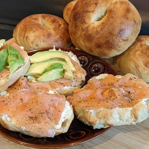 May include: A brown plate holds bagel halves topped with cream cheese, thin slices of pink smoked salmon sprinkled with dried dill, and fresh green avocado slices. Several whole, golden-brown toasted bagels are arranged behind the plate.