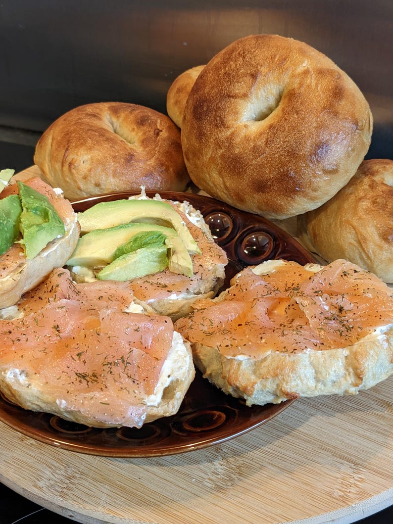 May include: A brown plate holds bagel halves topped with cream cheese, thin slices of pink smoked salmon sprinkled with dried dill, and fresh green avocado slices. Several whole, golden-brown toasted bagels are arranged behind the plate.